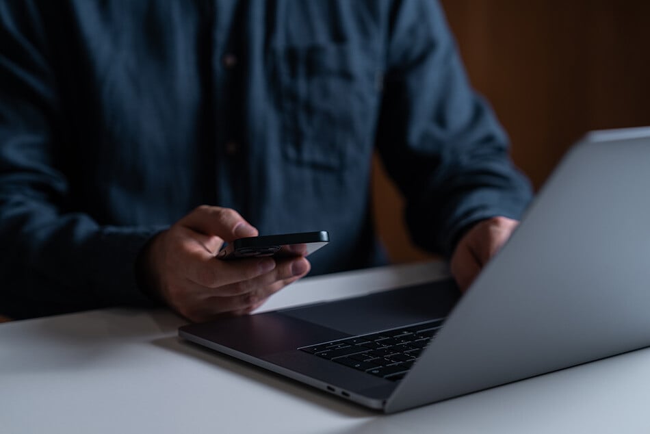 a-man-looks-at-his-phone-while-typing-on-his-laptop