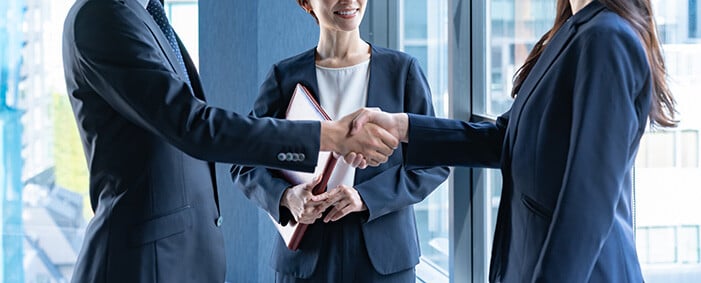 two people shake hands while a third person watches smiling.