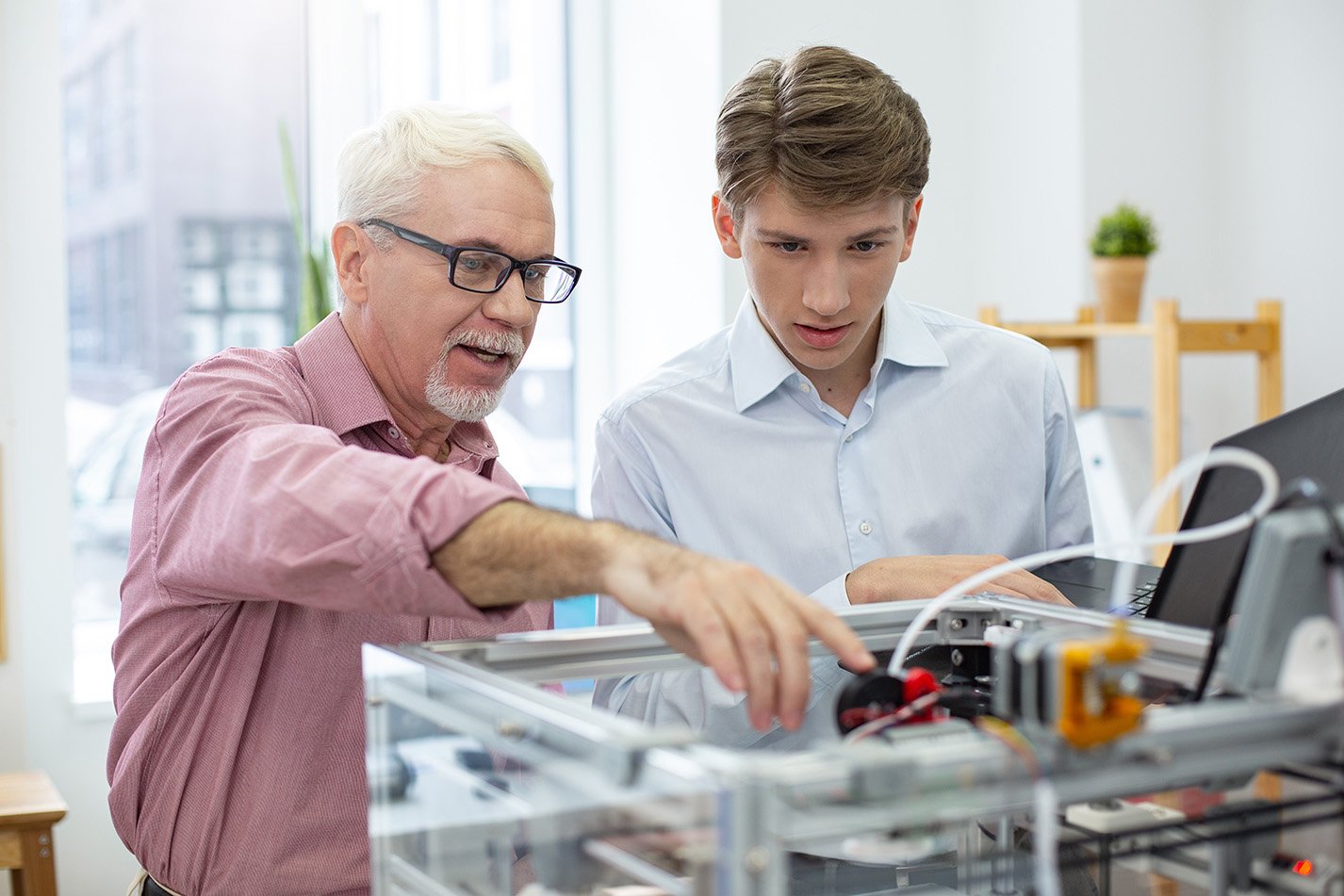 Experienced senior engineer instructing his young intern about 3D printers while pointing at the important parts of mechanism.