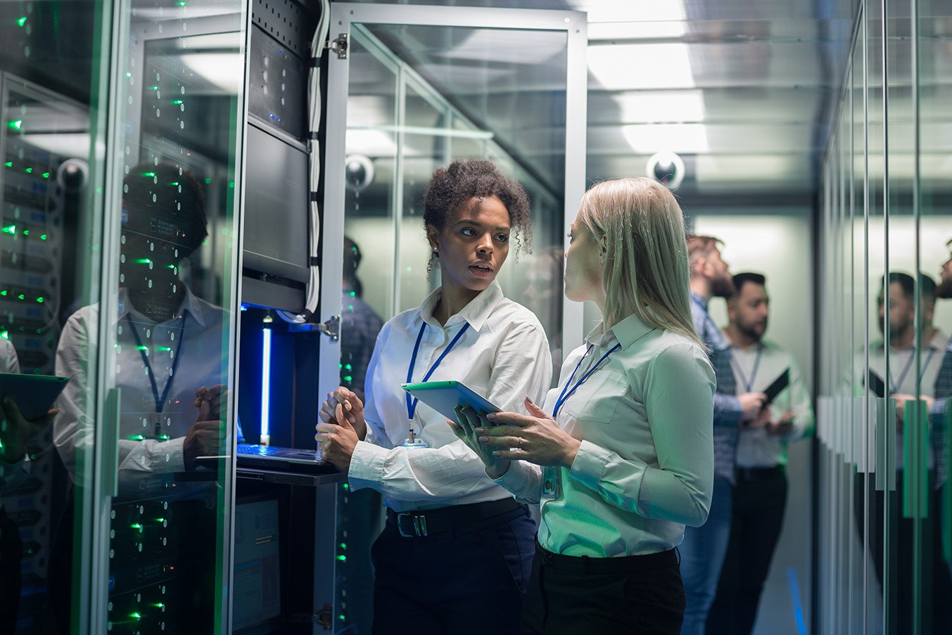 two women standing next to each other in a server room talking while one of them holds an ipad