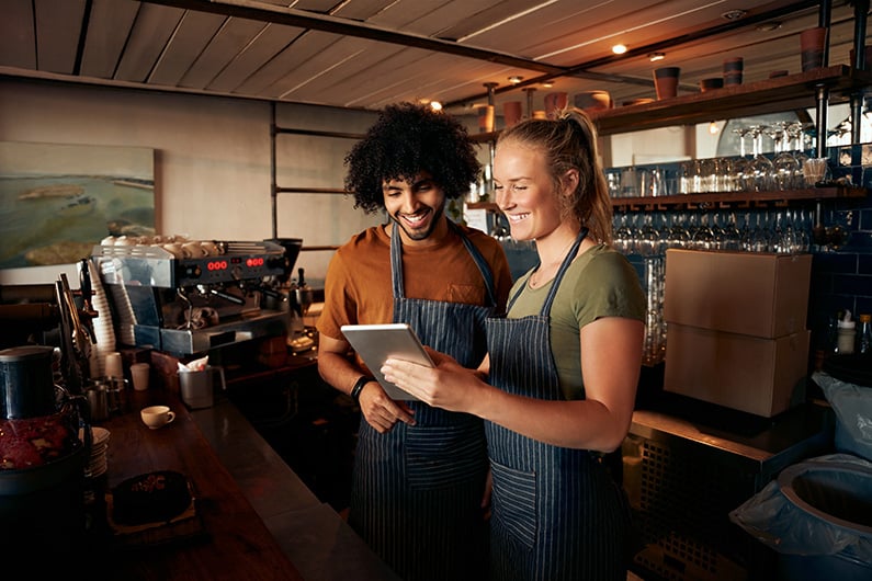 two restaurant workers smile and look at an ipad while behind a bar counter.