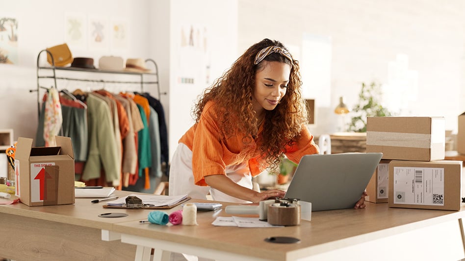 a female business owner at her desk with packaging materials looking at a laptop.