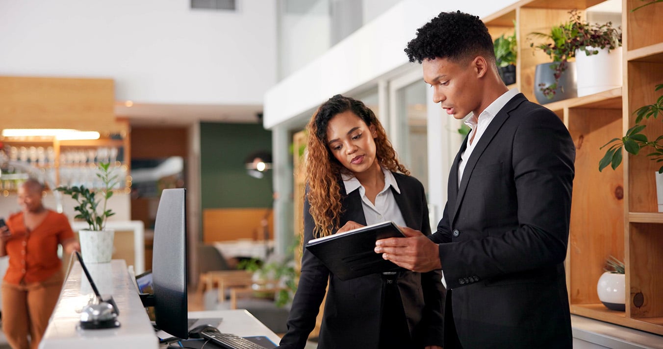 two hospitality workers one male one female standing at the front desk looking at a tablet.