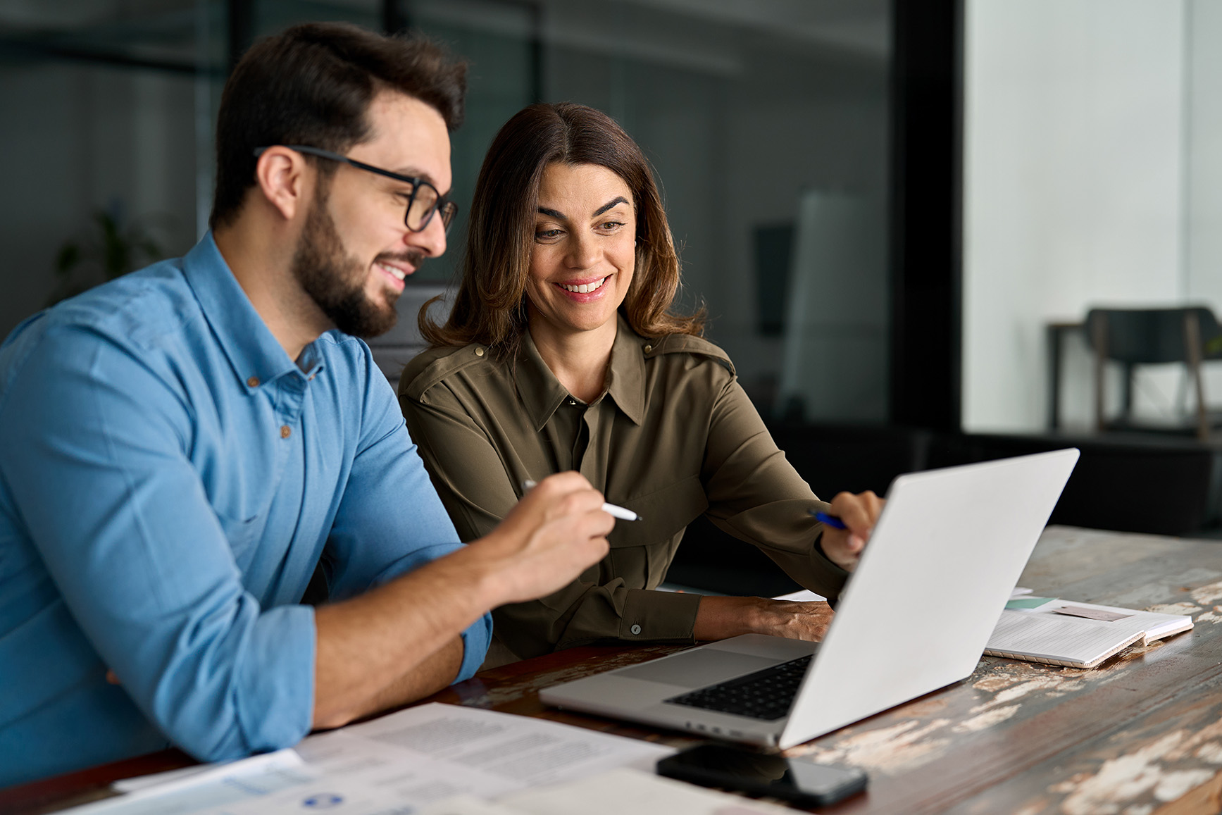 a man and a woman sit at a desk together and look at a laptop screen.