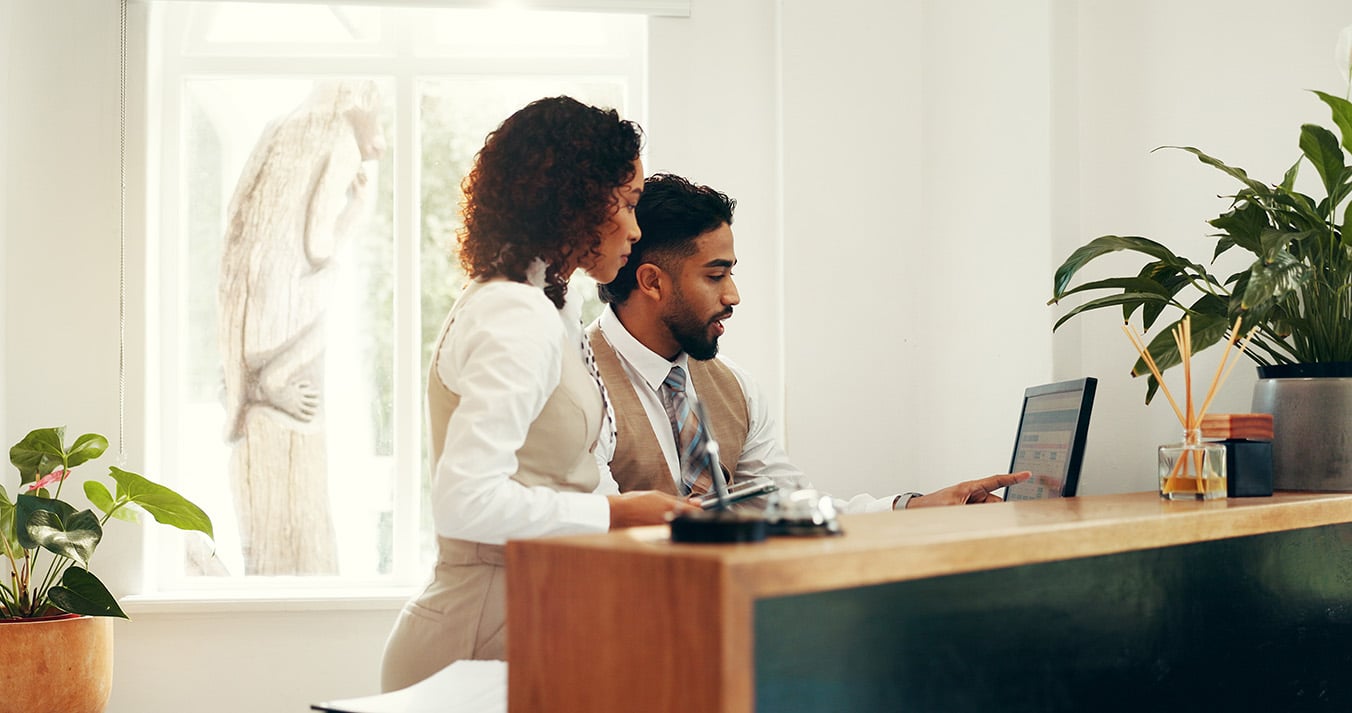Hotel, receptionist and colleagues with computer, research and collaboration for reservation on web. Online, man and woman with tech for registration info, teamwork and planning for schedule in motel.