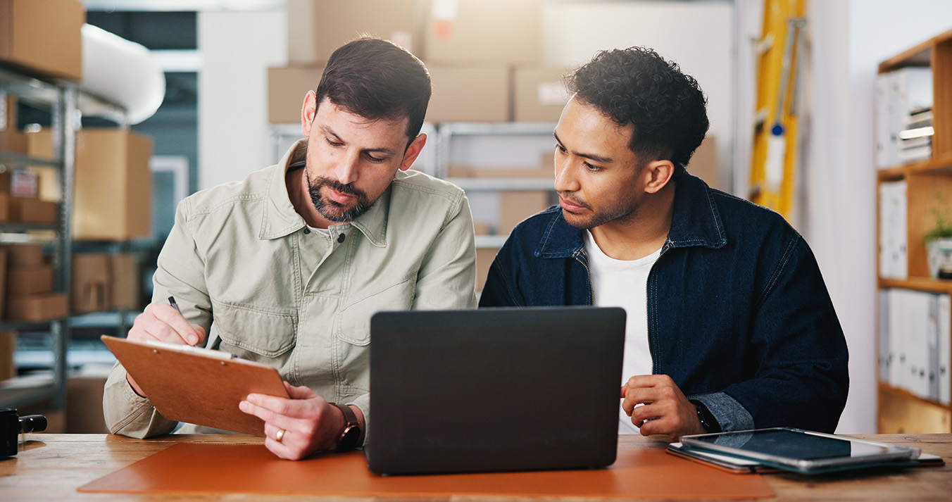 two men working at a small business looking over numbers on a clipboard and verifying with a laptop.