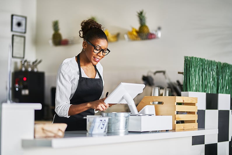 smiling-shop-assistant-using-pos-point-of-sale-terminal-to-put-in-order-from-notepad-at-restaurant-register