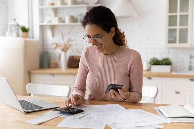 a-woman-sitting-at-her-kitchen-table-with-various-papers-and-her-laptop-typing-numbers-into-a-calculator
