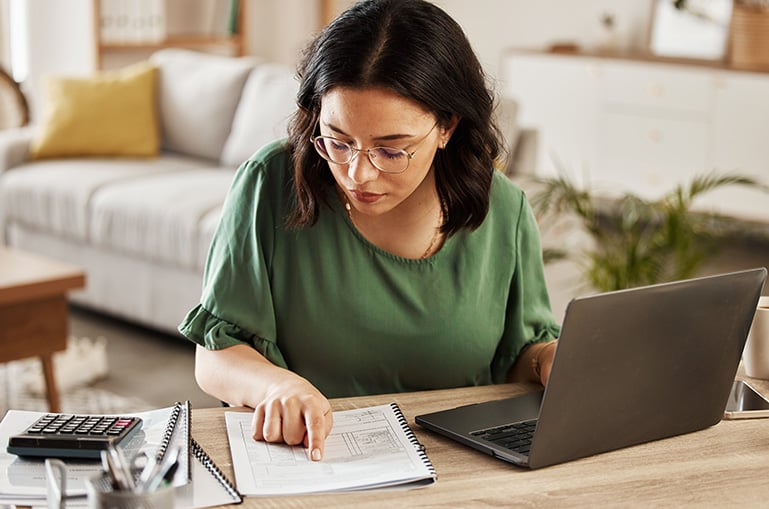a woman sits at a desk with her laptop and reads from a piece of paper