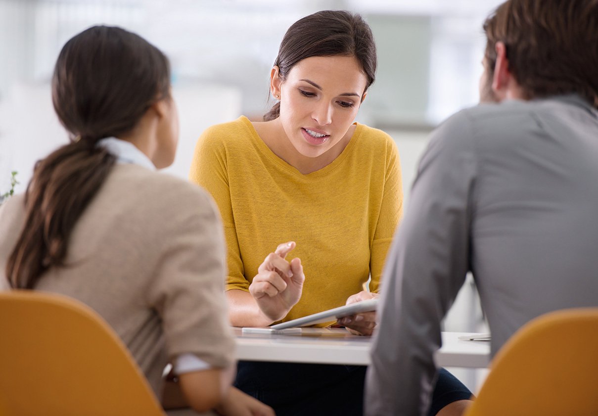 a woman sits across from a pair of employees and explains something to them that is on a piece of paper